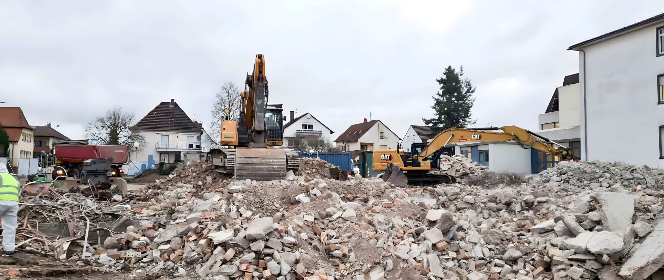 Auf einer städtischen Baustelle räumen zwei CAT-Bagger Bauschutt aus einem ehemaligen Gebäude. Im Hintergrund Wohnhäuser, ein Lkw und ein Mitarbeiter in Warnkleidung