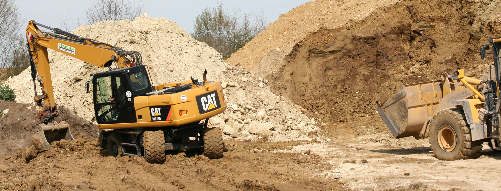 Zwei Radbagger, ein gelber CAT-Bagger und ein gelber Kürzl Radlader, heben Erde auf einer großflächigen Baustelle. Große Erdhügel und Bäume im Hintergrund.