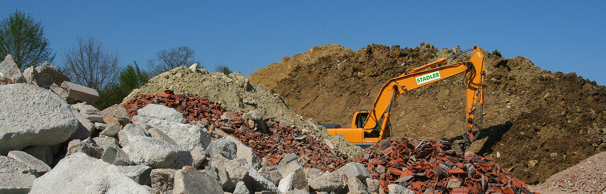 Bagger der Fa. Stadler bei Erdarbeiten auf einer Baustelle, im Vordergrund Bauschutt und Dachziegel